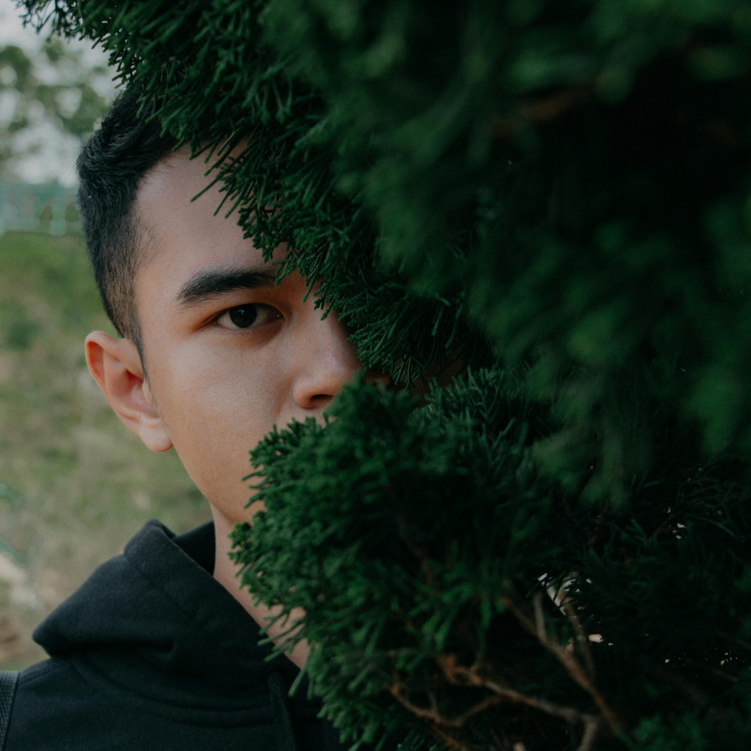 Close-up of a young man partially hidden behind green foliage, conveying mystery and curiosity.